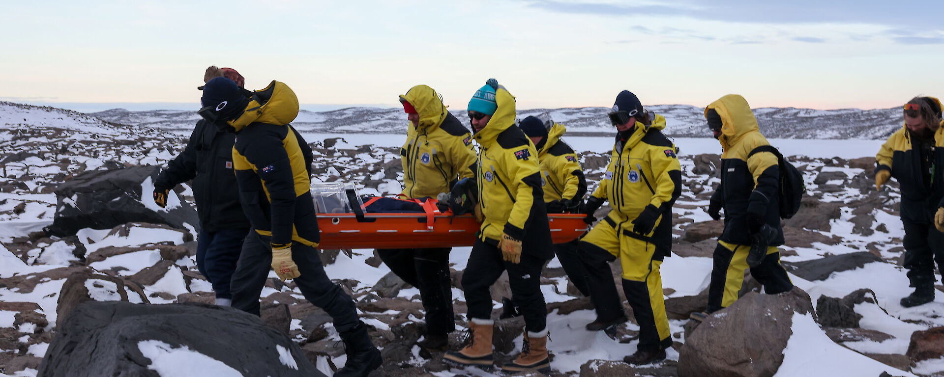 Six people in yellow carry a red stretcher across a rocky, snowy landscape