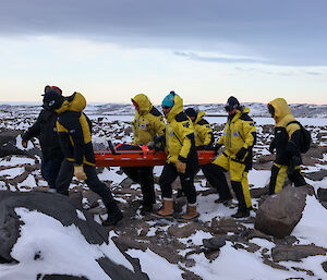 Six people in yellow carry a red stretcher across a rocky, snowy landscape