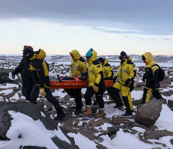 Six people in yellow carry a red stretcher across a rocky, snowy landscape
