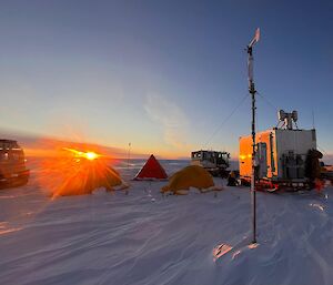 A radiating sun stoops the snow stricken horizon, luminating a remote camp of three tents, two vehicles and a transportable shelter. An audacious aerial weather station in the fore-ground.