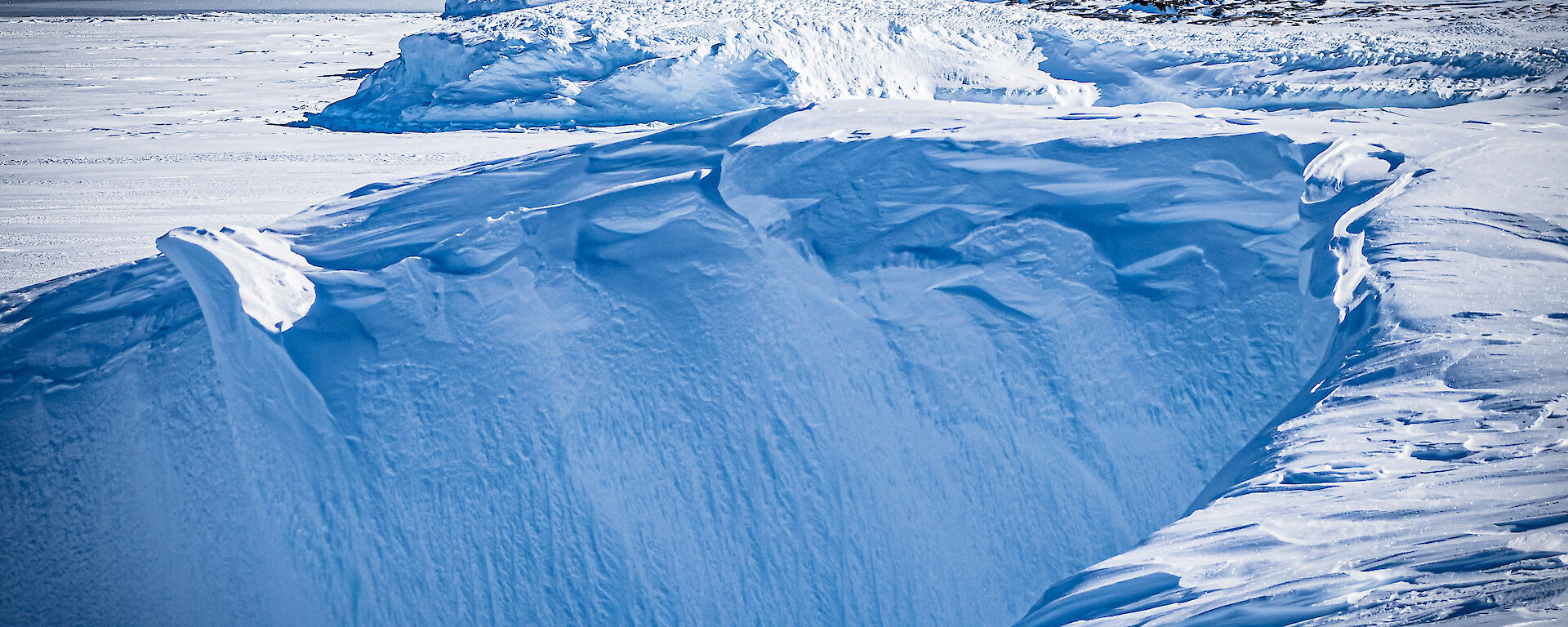 A landscape photo of foreground ice cliff billowed frozen with midground fast-ice edge into open ocean and a backdrop of icebergs