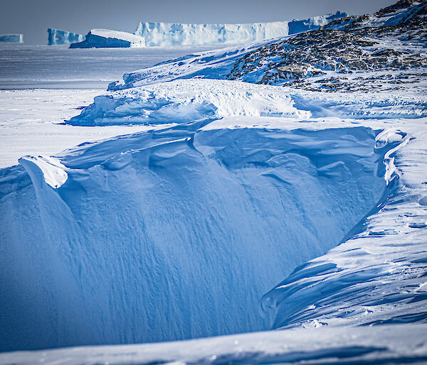 A landscape photo of foreground ice cliff billowed frozen with midground fast-ice edge into open ocean and a backdrop of icebergs