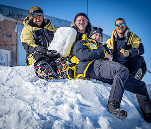 Four expeditioners sitting on a snow mound outside Wilkes hut, smiling, one holds a large block of snow. The backdrop includes a humorous 'Hilton' sign.