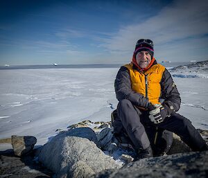 Dave buller sits on top a rock for a photograph, with a view of Shirley island to the right mid-ground and sea ice into fast ice edge to open water.