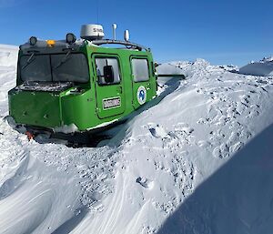 A Hagglund vehicle is partially buried in deep snow. The snow drifts arounds the vehicle with treads and lower half covered. Clear sky in the distant