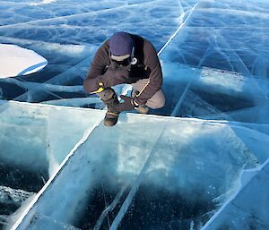 A man in dark grey crouches on a frozen lake which is clear, frozen to 1.5 metres