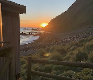 Sun setting over the sea peaking around side of a mountain that drops down to the coast. In the foreground, side of a hut with fenceline.