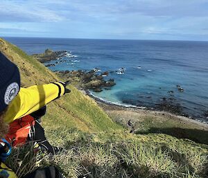 Man points down steep glassy slope to coast line and hut in the distance
