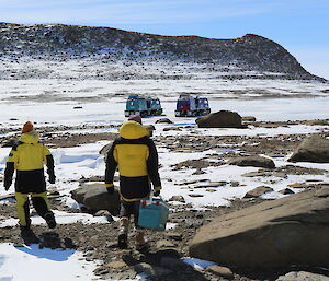 Three people in yellow and black jackets walk towards two oversnow vehicles in the distance, with a rocky, snow-covered mountain in the background.
