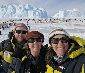 Three people taking a selfie in front of a distant emperor penguin colony.