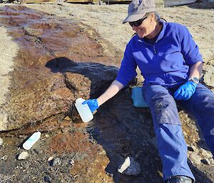 A woman sitting beside a meltwater stream running across a large slab of rock in front of a building. The woman is collecting water from the stream in a small bottle.
