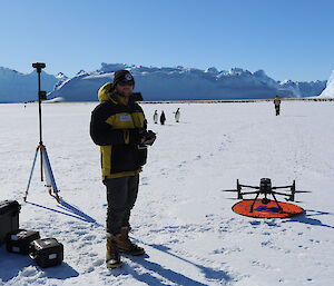 A man in a puffy yellow jacket standing beside a drone sitting on an orange circle, some distance from an emperor penguin colony on ice, with icebergs in the background