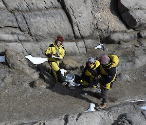 The photographer looks down on three people in a gully between two rocky slopes. The three people are standing and kneeling beside a white bucket in which they're collecting soil samples.