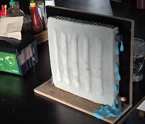 Cake decorated to look like a wall hydronics radiator, standing on it's end, covered in cream icing and with valves and water droplets made of icing