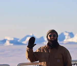 Jono Newman waving from the deck of a ship, with icebergs in background.