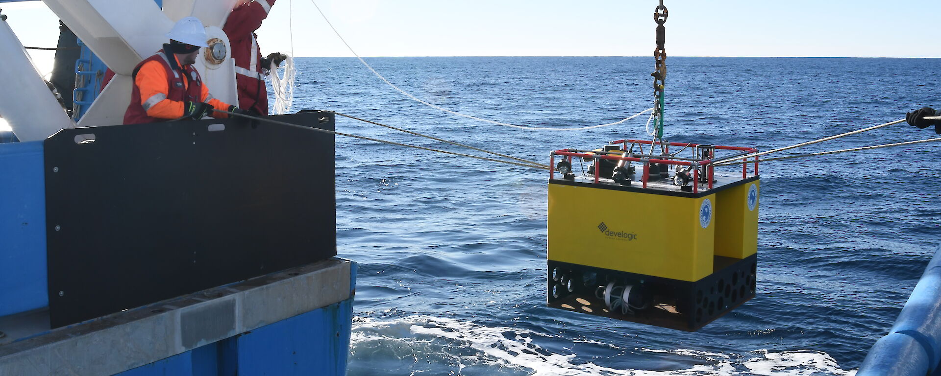A square yellow instrument being lowered in to the ocean from a chain, with people each side on the ship's deck steadying it with ropes.