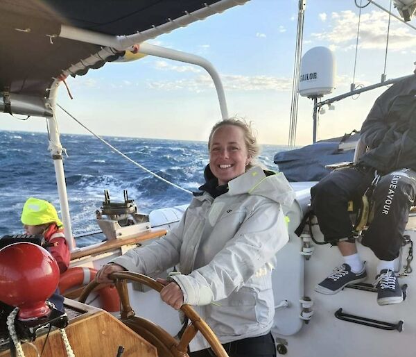A young woman, smiling, wearing a white jacket, standing on an older style sailing vessel, with the large ships wheel. Background of photo shows the open ocean.