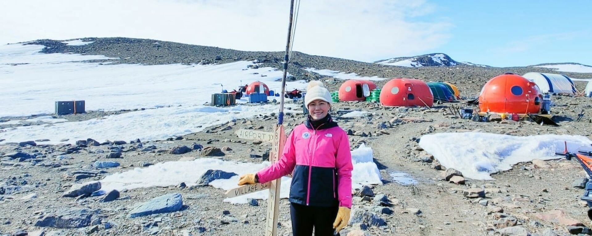 Image of Jacqui at Denman Terrestrial Campaign, Bunger Hills. Photo displays Jacqui standing with Australian flag in foreground and the camp in the background. Rocky and snowy landscape fill the distance.