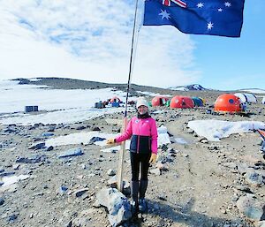 Image of Jacqui at Denman Terrestrial Campaign, Bunger Hills. Photo displays Jacqui standing with Australian flag in foreground and the camp in the background. Rocky and snowy landscape fill the distance.