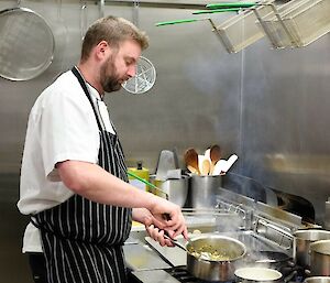 Image of John working in the kitchen. John is in the foreground stiring food in a pot.