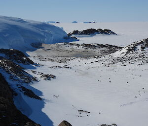 Looking down a valley of snow, containing the emperor penguin colony, with rocky ridges down either side, and Taylor Glacier on the right hand side. Sea ice and icebergs in the distance