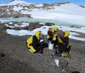 Cath, Kathryn and Gwil kneeling on grey soil and rocks. Cath is digging with a trowel and Kathryn and Gwil are preparing the containers for the soil sample to go into. There are some hills in the background, some snow drifts and a small frozen lake. It is a sunny blue-sky day