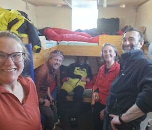 Selfie taken by Cath of the team crowded into the tiny Colbeck Hut, with bunk beds on the left and rear walls.  From left to right, Cath, Gwil, Daniel, Jason, Kathryn, and Jason. Kathryn has a handful of dice, ready to roll in a game of Farkle