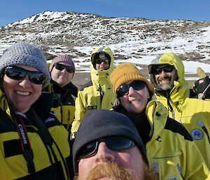 Selfie of the whole team taken by Cath, with Jason in the foreground, and from left to right Cath, Gwil, Daniel, Kathryn and Jason all wearing yellow jackets, beanies and sunglasses. A rocky hill covered with patchy snow is in the background. It is a blue-sky sunny day