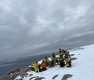 Eight expeditioners in yellow survival gear pose for a photo. The background features the ocean and ice bergs.