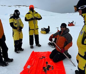 Field Training Officer kneels on tarp with sea ice drill demonstrating its use. Expeditioners in survival gear watch on for the demonstration. The background is grey sky with snow covering the ground.