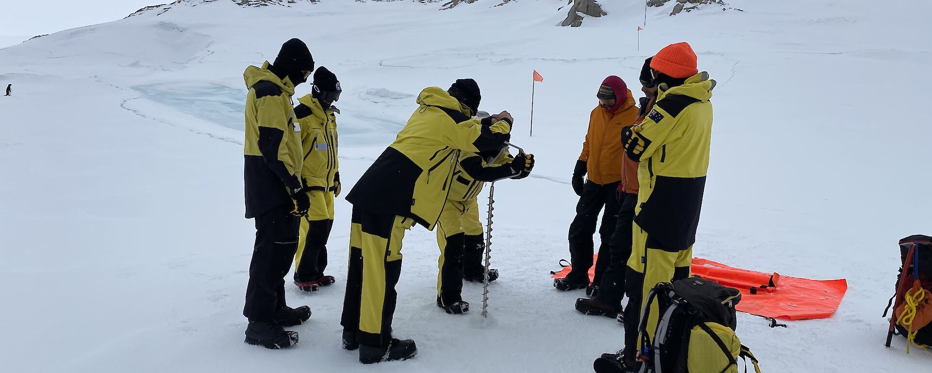 Image of seven expeditioners in survival clothing learning to drill the sea ice. Survival Packs are scattered in the foreground. Small rocky hills are seen in the background.