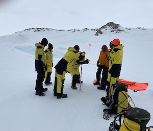 Image of seven expeditioners in survival clothing learning to drill the sea ice. Survival Packs are scattered in the foreground. Small rocky hills are seen in the background.