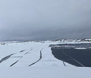 Image features the sea ice breaking away from the edges of the land. Hills covered in snow line the horizon in the background. The sky is grey and moody. The image feels cold.