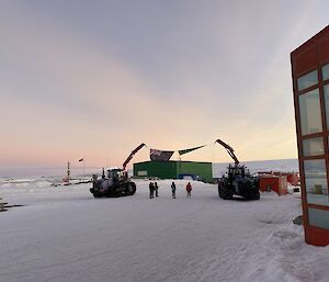 Two tractors with cranes extended. Flags hung between them simulating an archway. Sunrise on the horizon. Snow on the ground. Red shed to the right of the image. Four people stand under the archway. Resembles a closing scene to a movie.