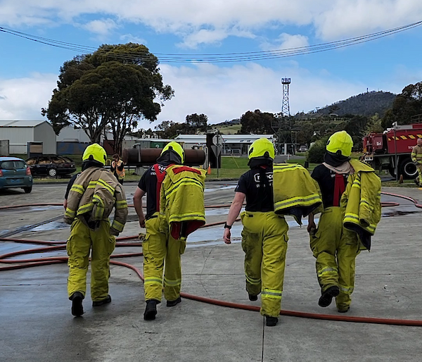 Four people walk in a line abrest away from the camera. The individuals are wearing fire fighting suits with their jackets thrown over their shoulder. The ground is concrete with fire hoses scattered across the mid-ground. The sky is blue with clouds. A fire truck can be partially seen in the mid-ground.