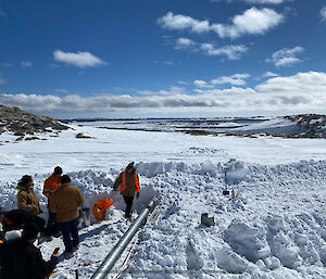 Image of plumbers in high vis gear working in the foreground on broken pipework. Snow covers the ground and site services in the foreground. The background features Newcomb Bay and the antarctic landscape.