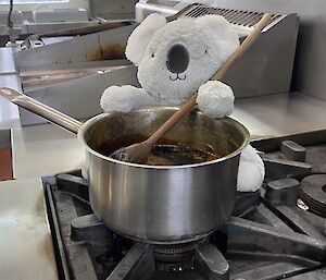 Toy koala sits on the stove and holds a wooden spoon in a saucepan
