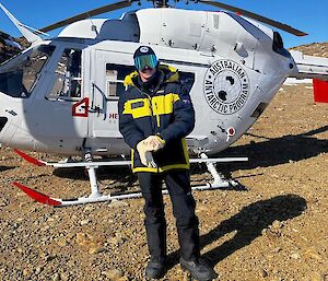 A man in outdoor gear and goggle stands in front of a white helicopter