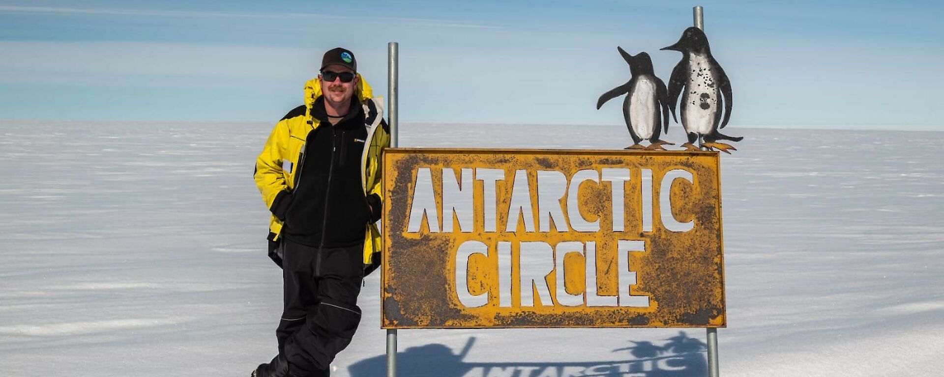 Expeditioner Jacob leaning against the Antarctic Circle sign. The background shows vast flat white snow into the distance with a clear blue sky to the horizon.