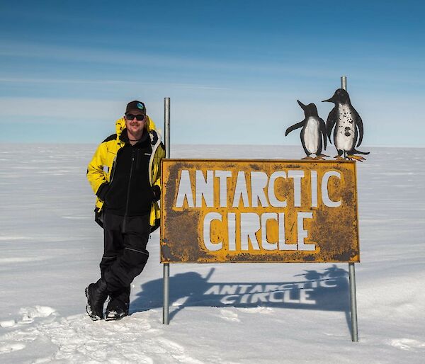 Expeditioner Jacob leaning against the Antarctic Circle sign. The background shows vast flat white snow into the distance with a clear blue sky to the horizon.