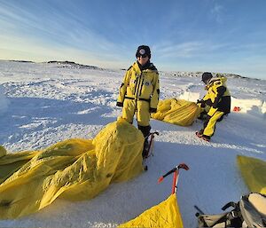 Image displays Expeditioner in Yellow Survival suit rolling out yellow bivvy bag on the snow.