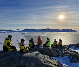 Image of 6 expeditioners sitting on rocks near a cliff. The image highlights the scenery. In the mid-ground is the ocean meeting the broken sea ice. The background features snowy hills and the sun in the middle of the sky.