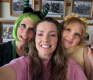 Three women dressed up in a green wig, pink eye glitter and a gold head piece