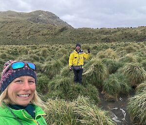 A woman with a beanie on stands in tussock grass with a man in yellow in the background