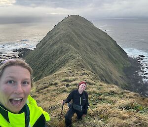 Two women stand on a grassy ridge with rocks and sea in the background