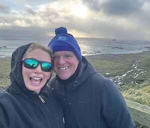 Two smiling people stand on a wooden platform with sea and grassy hill behind them