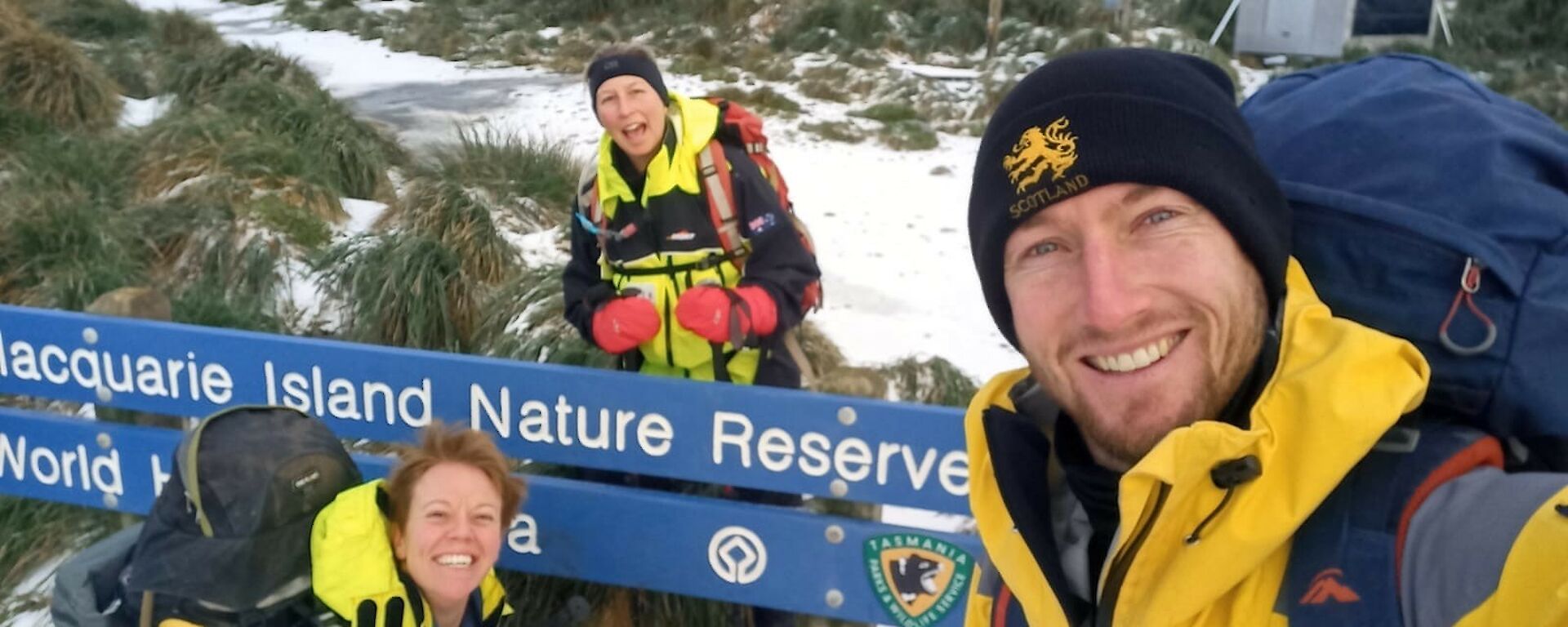 A man and two women with snow around, near the Macquarie Island Nature Reserve sign