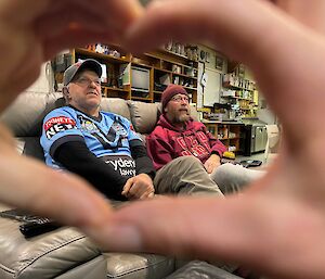 A person making a heart shape with their hands and inside are two men sitting on a couch