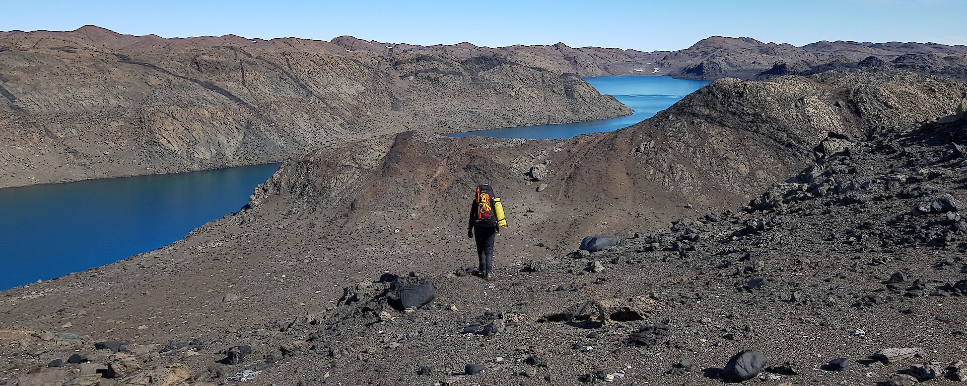 A person walking through a brown landscape of rocky hills with a blue lake dividing them.