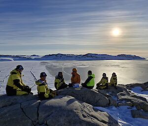 Seven Expeditioners sit along a rock with sea ice in the mid-ground. The background is a light blue sky with a bright sun.